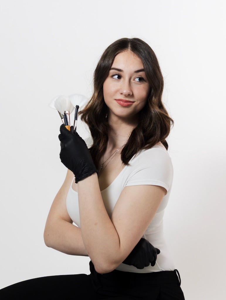 Paulina, licensed esthetician at PK Beauty in Trumbull, Connecticut, in professional attire with black gloves, holding skincare fan brushes on a white backdrop